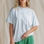 Woman wears an oversized pale blue t-shirt with short sleeves, a round neckline, and small "Foxwood Athletics" chest print. Styled casually with black leggings.