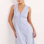 A woman modeling a sleeveless light blue and white gingham maxi dress with a V-neckline and front button detail, standing against a plain white wall.