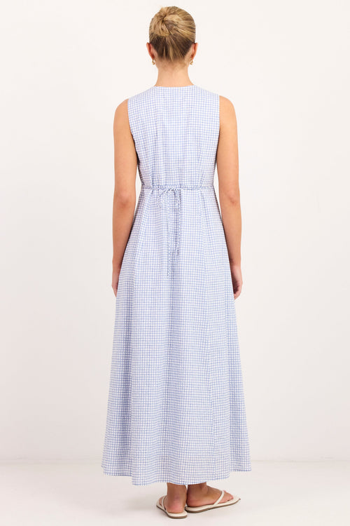 A woman viewed from behind wearing a sleeveless light blue and white gingham maxi dress with a tie waist, standing against a plain white studio background.