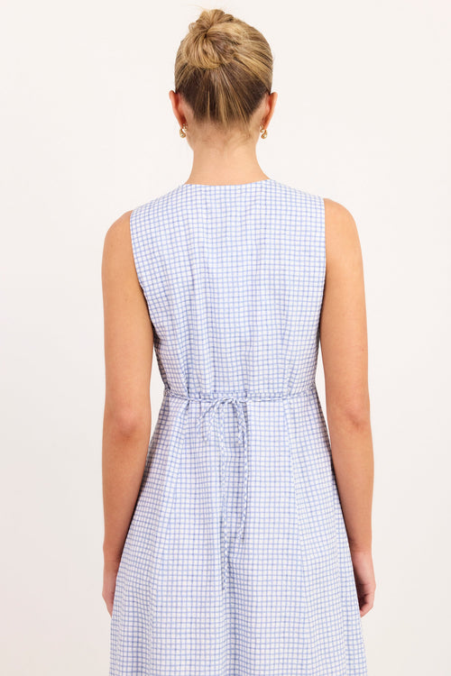 A woman viewed from the back wearing a sleeveless light blue and white gingham dress with a tie waist, set against a plain off-white background.