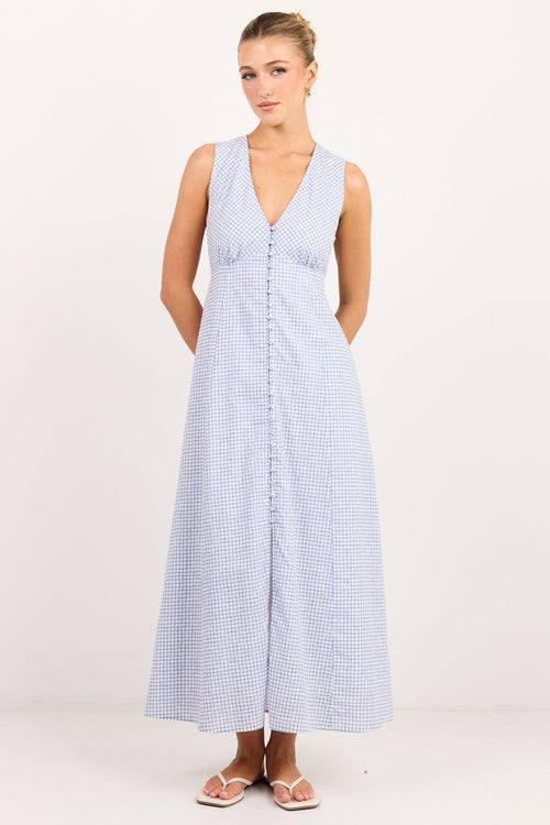A woman modeling a sleeveless light blue and white gingham maxi dress with a V-neckline and front button detail, standing against a plain white studio background.