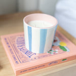 A pastel pink and white striped candle jar sits on top of a pink book on a light wood surface, with a softly blurred home interior in the background.