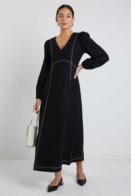 Woman wearing a black maxi dress with balloon sleeves and contrast stitching, styled with black flats and sunglasses