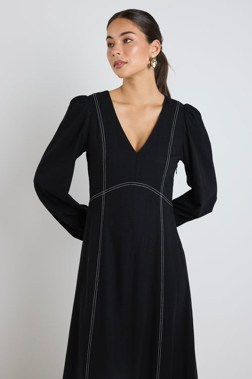 Woman wearing a black maxi dress with balloon sleeves and contrast stitching, styled with black flats and sunglasses