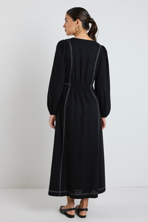 Woman wearing a black maxi dress with balloon sleeves and contrast stitching, styled with black flats and sunglasses