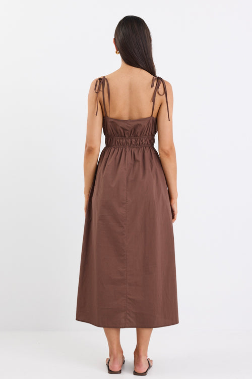 A brown cotton sundress featuring adjustable straps, a tie-front detail, an elasticated waist for a flattering fit, and side pockets. The dress is ankle-length with a relaxed fit.