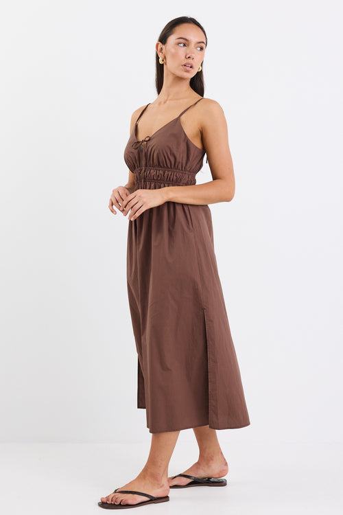 A brown cotton sundress featuring adjustable straps, a tie-front detail, an elasticated waist for a flattering fit, and side pockets. The dress is ankle-length with a relaxed fit.
