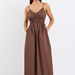 A brown cotton sundress featuring adjustable straps, a tie-front detail, an elasticated waist for a flattering fit, and side pockets. The dress is ankle-length with a relaxed fit.