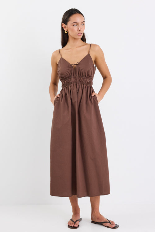 A brown cotton sundress featuring adjustable straps, a tie-front detail, an elasticated waist for a flattering fit, and side pockets. The dress is ankle-length with a relaxed fit.