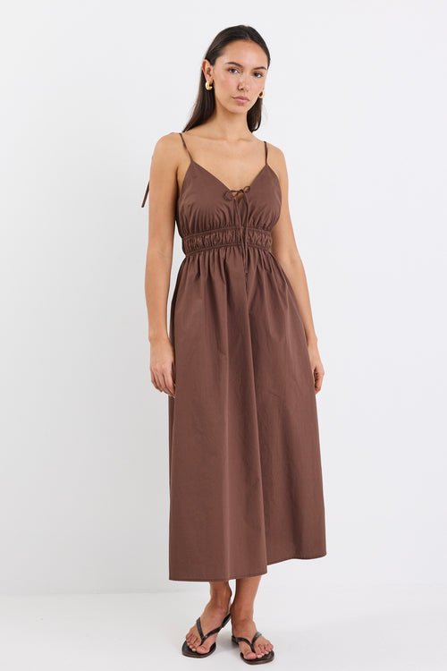 A brown cotton sundress featuring adjustable straps, a tie-front detail, an elasticated waist for a flattering fit, and side pockets. The dress is ankle-length with a relaxed fit.