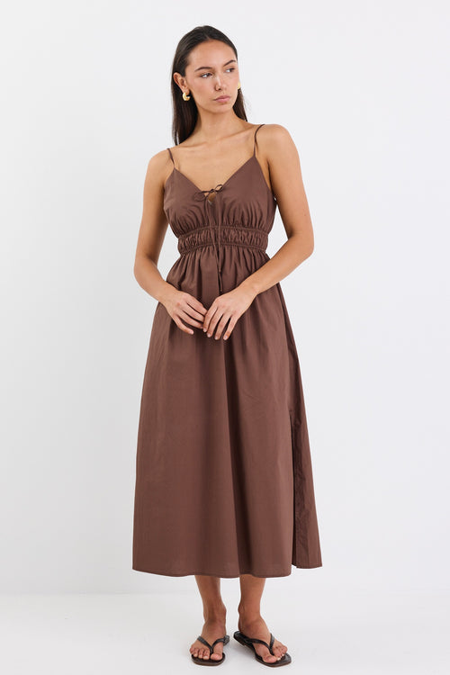 A brown cotton sundress featuring adjustable straps, a tie-front detail, an elasticated waist for a flattering fit, and side pockets. The dress is ankle-length with a relaxed fit.