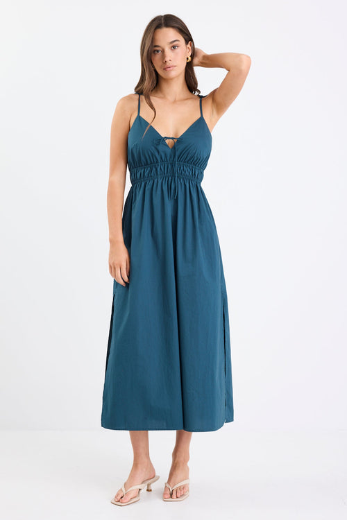 Woman wearing a petrol blue strappy cotton midi dress with a tie detail at the bust, gathered waist, and side splits.