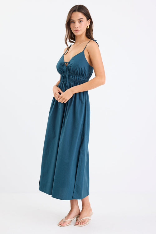 Woman wearing a petrol blue strappy cotton midi dress with a tie detail at the bust, gathered waist, and side splits.