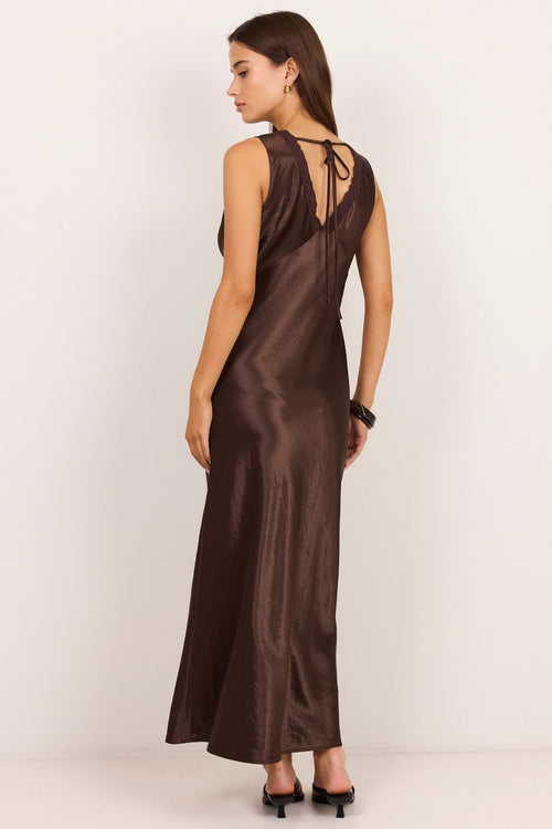 A woman modeling a dark brown satin slip dress from the back, featuring a deep V-neckline with tie-back straps, set against a plain off-white studio background.
