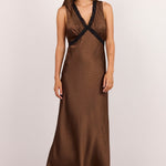 A woman modeling a sleeveless brown maxi dress with a textured pattern and black lace trim at the V-neckline, standing against a plain off-white studio background.
