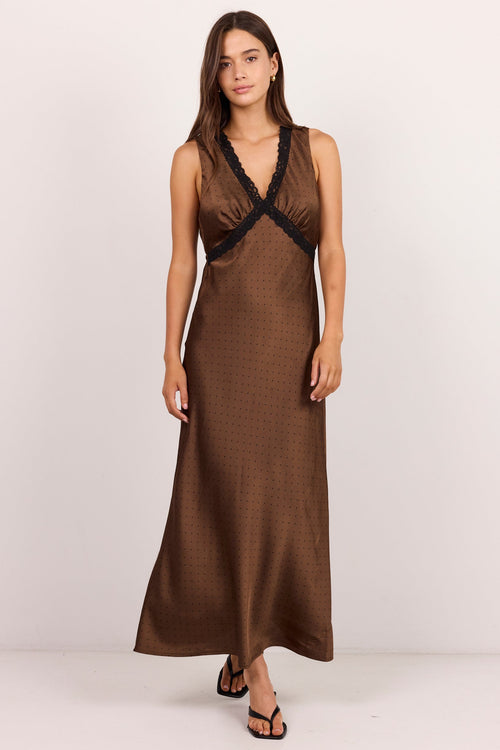 A woman modeling a sleeveless brown maxi dress with a textured pattern and black lace trim at the V-neckline, standing against a plain off-white studio background.