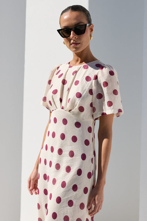 A woman modeling a cream-colored midi dress with dark red polka dots, styled with black sunglasses, standing in a bright white architectural setting with strong shadows.