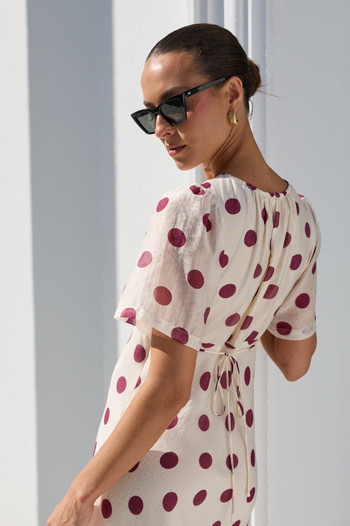 A woman wearing a cream-colored midi dress with dark red polka dots, styled with black sunglasses, standing in a bright, modern white room.