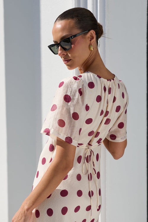 A woman wearing a cream-colored midi dress with dark red polka dots, styled with black sunglasses, standing in a bright, modern white room.