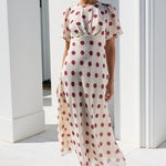 A woman wearing a cream midi dress with red polka dots and sunglasses, walking outdoors on a white tiled patio in front of a white building with columns.