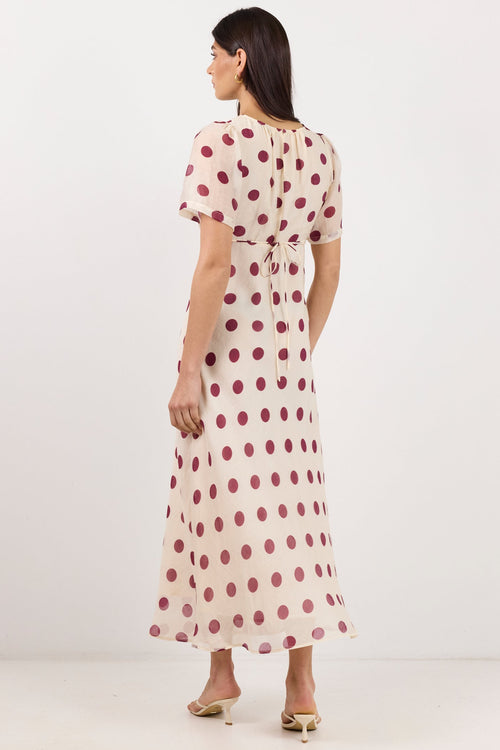 A woman is shown from the back wearing a short-sleeved midi dress with a beige base and dark red polka dots, paired with beige high-heeled sandals, set against a plain white studio background.