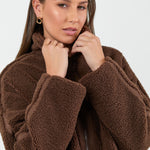 Woman wearing a chocolate brown sherpa fleece zip jacket and blue jean against a white background