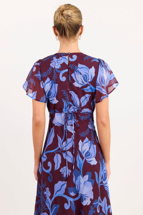 A woman viewed from behind wearing a short-sleeve wrap-style dress with a blue floral print on a deep burgundy background, set against a plain off-white studio backdrop.