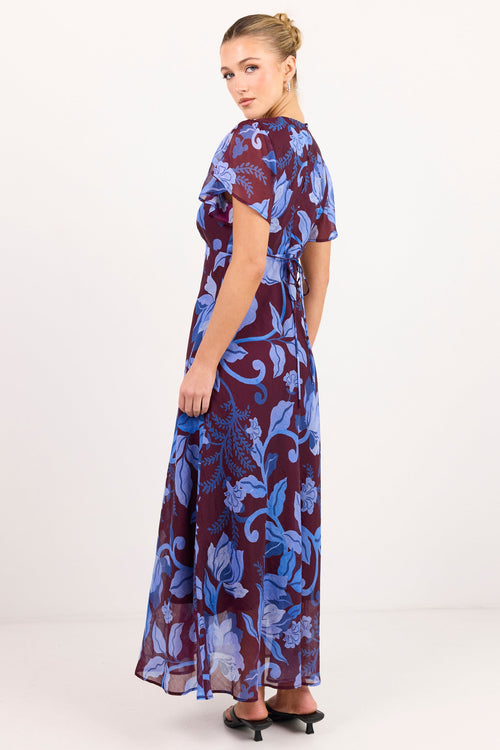 A woman modeling a short-sleeved maxi dress with a blue and purple floral paisley pattern, photographed from the back against a plain white studio background.