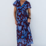 A woman modeling a short-sleeve maxi dress with a blue and burgundy floral pattern, standing against a minimalist white background with a circular architectural element.