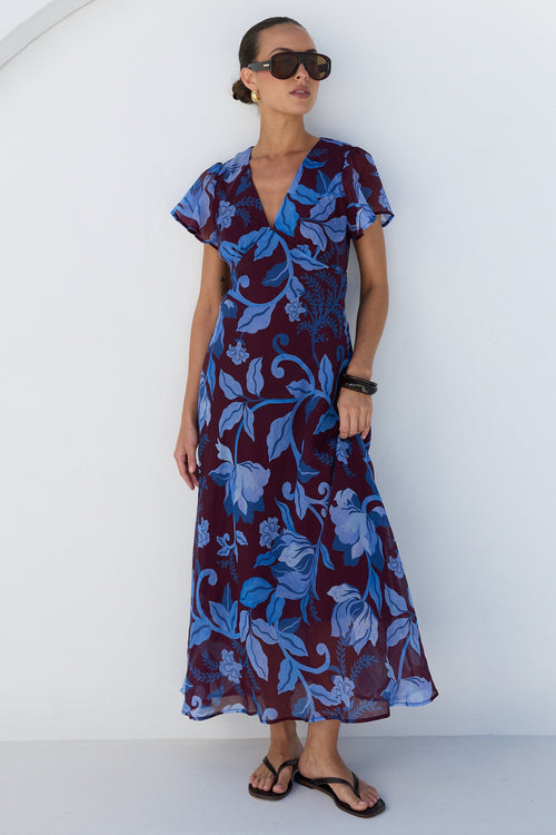 A woman modeling a short-sleeve wrap maxi dress with a blue and burgundy floral pattern, styled with black sunglasses and black sandals, standing against a white wall with an arched architectural detail.