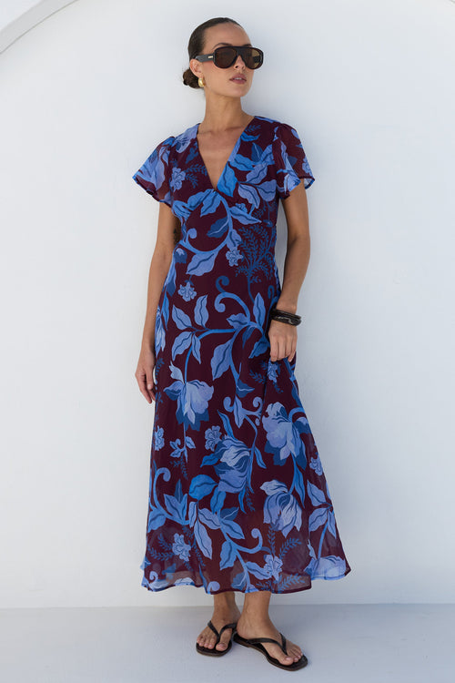 A woman modeling a short-sleeve maxi dress with a blue and burgundy floral pattern, styled with black sunglasses and black sandals, standing against a white wall with an arched background