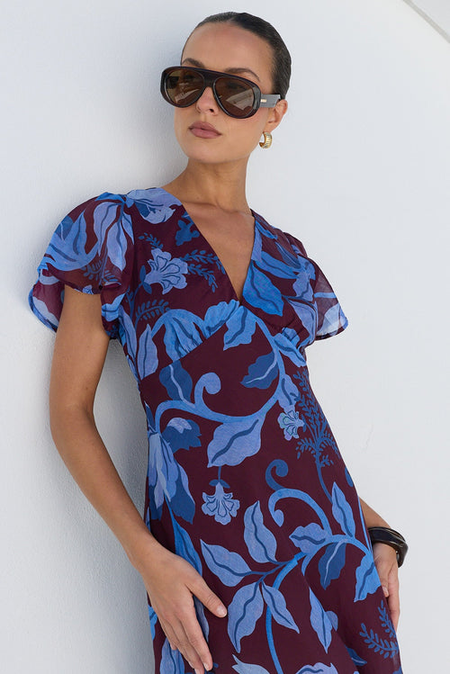 A woman wearing a dark burgundy maxi dress with a blue floral print, styled with sunglasses, standing against a white arched wall.