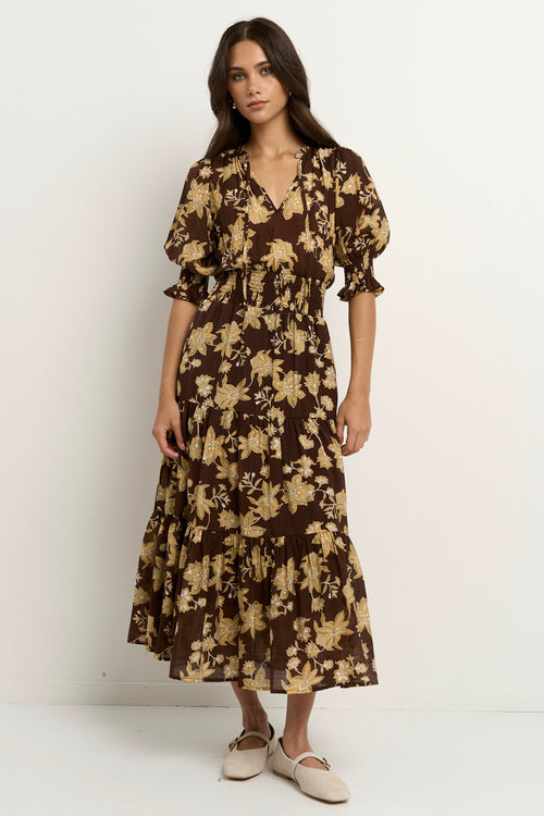 Woman wearing a brown floral midi dress with puff sleeves, a shirred waist, and a tiered skirt.