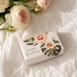A wrapped gift box with floral artwork and the words 'Happy Birthday' lies on white fabric next to a bouquet of pale blush and cream roses with green leaves.