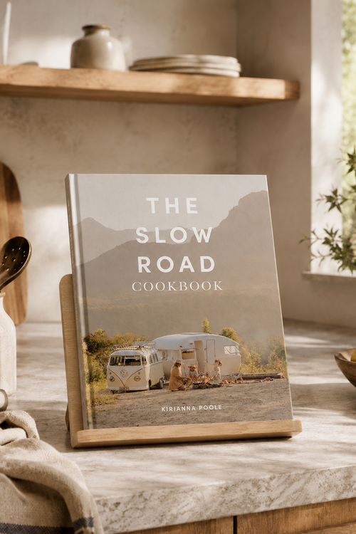 The Slow Road Cookbook by Kisianna Roole standing on a wooden stand on a stone countertop in a warm, rustic kitchen setting.