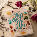 A book titled "The Art of Joy" by Jen Sievers lies on a white marble tray, styled with two purple tulips in a glass vase on a textured cream fabric background.