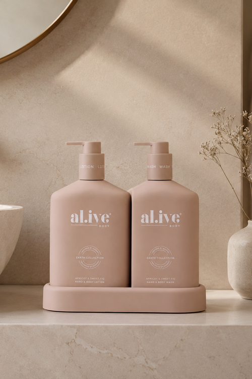 Two aLive Body wash bottles in a dusty pink color, sitting on a matching pink holder on a beige marble bathroom counter next to a white vessel sink and a vase with dried flowers.
