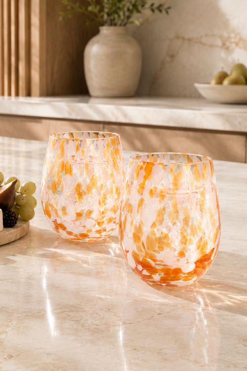Two stemless wine glasses with an orange and white marbled pattern, set on a light marble kitchen counter next to a charcuterie board with cheese, grapes, and crackers.