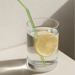 A clear glass tumbler with a green curved straw, filled with water and a lemon slice, set on a light beige surface against a textured beige wall with strong diagonal shadows.