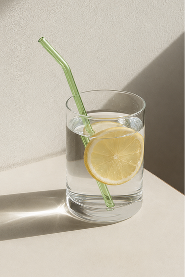 A clear glass tumbler with a green curved straw, filled with water and a lemon slice, set on a light beige surface against a textured beige wall with strong diagonal shadows.