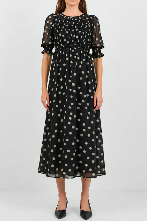 A woman modeling a black midi dress with a white polka dot pattern, featuring short puff sleeves and a gathered waist, set against a plain light gray studio background.