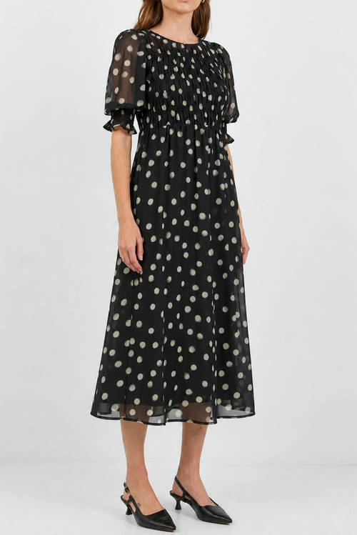 A woman modeling a black midi dress with a white polka dot pattern, featuring short puff sleeves and a gathered waist, paired with black pointed-toe heels against a light gray studio background.