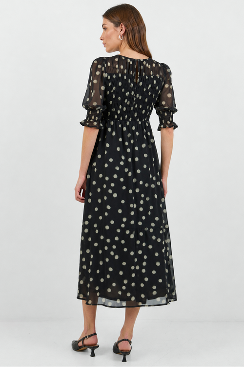 A woman modeling a black midi dress with a white polka dot pattern, shown from the back against a light gray studio background.