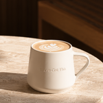 A white ceramic coffee mug with a handle, filled with a latte and resting on a round stone table in warm sunlight.