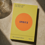 A book titled "SPRITZ" by Spritz, featuring a minimalist yellow cover with a large orange circle and blue text, photographed on a textured beige stone surface with dramatic shadows.