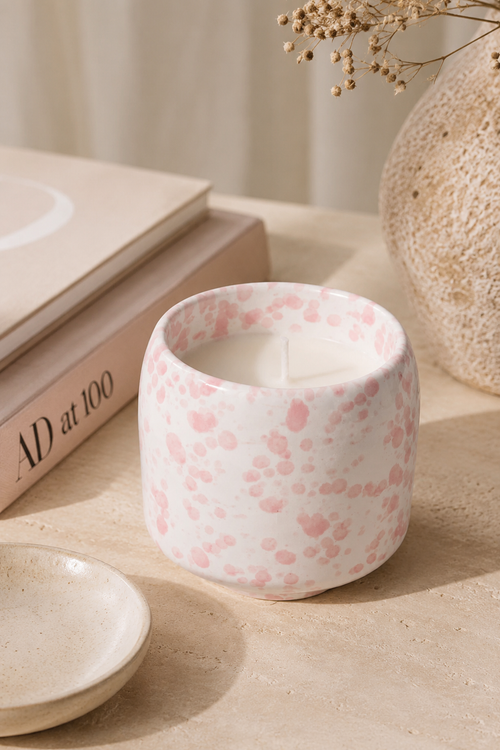 A white ceramic candle jar with a pink speckled pattern, styled on a beige surface next to a small dish, books, and a textured vase.