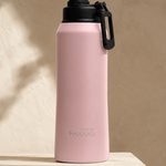 A pink insulated water bottle with a black lid and carrying loop, standing on a textured beige stone pedestal against a beige wall with soft leaf shadows.