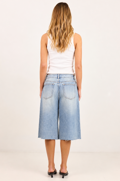 A woman viewed from behind wearing light-wash blue denim culottes, a white tank top, and black heels against a plain white studio background.