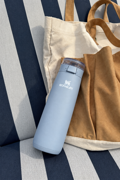 A light blue Stanley travel tumbler with a clear lid resting on a navy and white striped cushion next to a beige canvas tote bag with tan handles.
