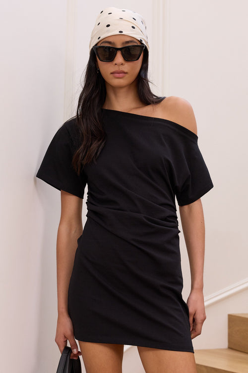 A woman models a black off-the-shoulder mini dress with gathered side detailing, styled with a black and white polka dot bandana, black sunglasses, and a black handbag, standing on wooden stairs against a white wall.